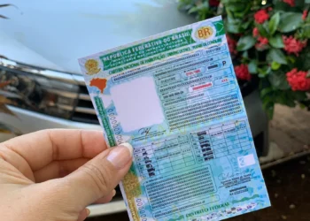 Carteira Nacional de Habilitação (CNH) sendo segurada por uma mão, com o carro visível ao fundo. Imagem: Detran