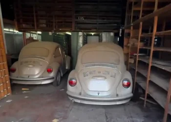 Dois Volkswagen Fusca abandonados e cobertos de poeira dentro de um antigo depósito com prateleiras vazias.