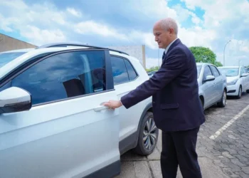 "Idoso vestido formalmente abrindo a porta de um carro branco em estacionamento."