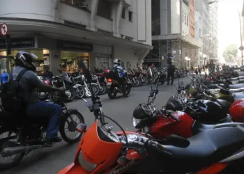 Rua movimentada com diversas motocicletas estacionadas e motociclistas circulando.