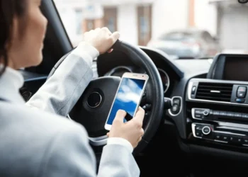 Mulher segurando um smartphone com uma mão enquanto dirige, com a outra mão no volante.