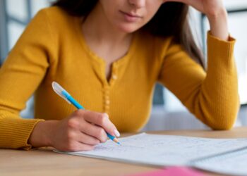 Mulher concentrada escrevendo e estudando em seu caderno, com caneta azul em uma mesa de trabalho.