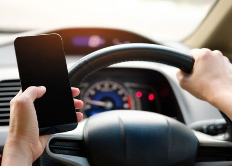 Mãos segurando volante do carro e celular simultaneamente.