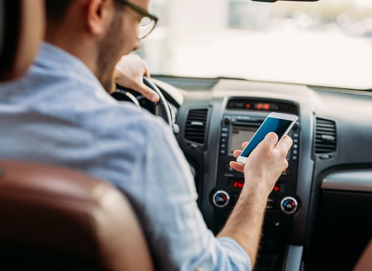Homem dirigindo carro enquanto segura celular com a mão direita.