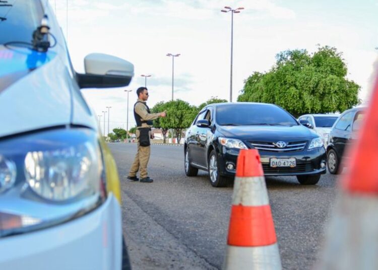 Agente de trânsito realizando uma abordagem de blitz a motorista em uma estrada, com cones de sinalização visíveis.