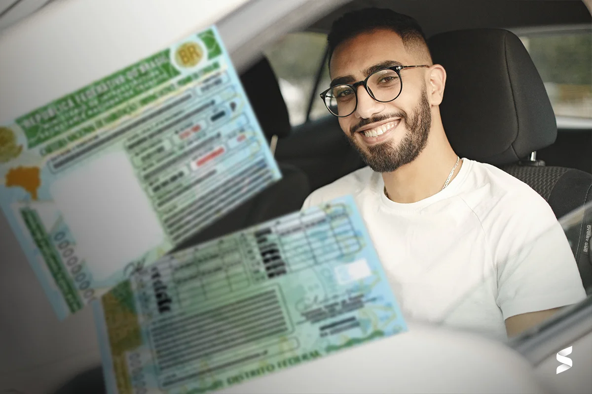 Homem sorrindo dentro de um carro enquanto segura sua Carteira Nacional de Habilitação (CNH), com foco no documento.