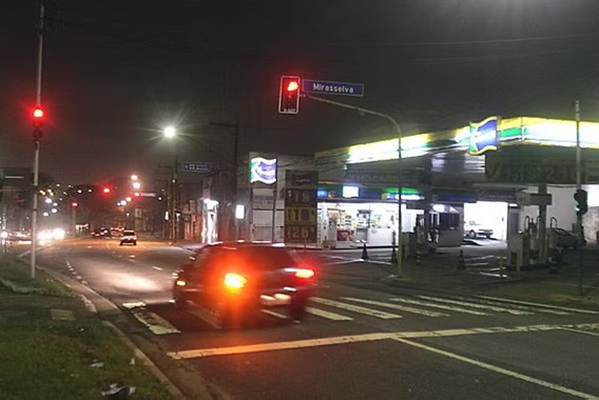 Veículo avança sinal vermelho em cruzamento urbano à noite, próximo a posto de combustível.