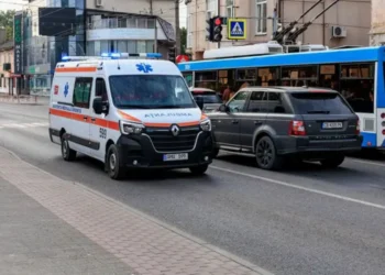 Ambulância se aproximando em uma rua movimentada, com carros bloqueando parcialmente a passagem.