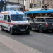Ambulância se aproximando em uma rua movimentada, com carros bloqueando parcialmente a passagem.