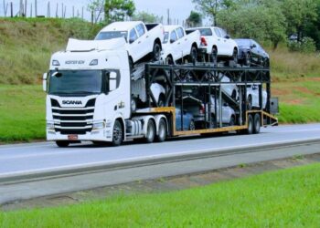 Caminhão cegonha carregando carros e caminhonetes brancas em plataforma elevada, trafegando pela estrada.