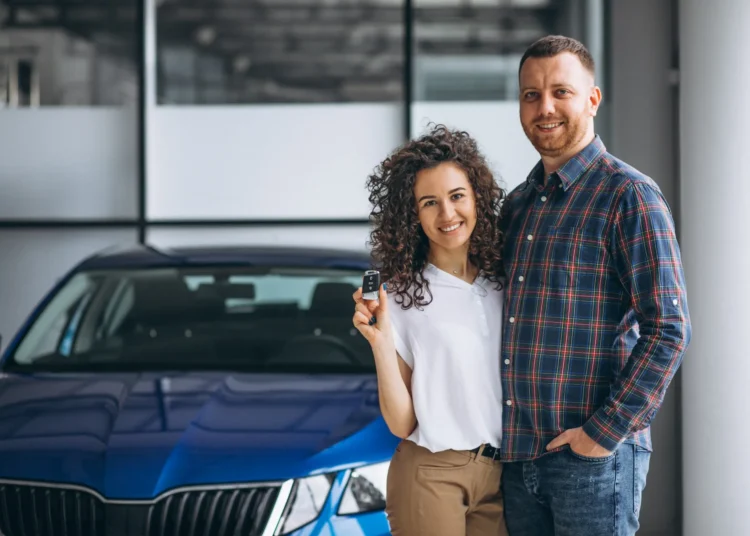 Casal jovem feliz em showroom de carros com chave de carro, aproveitando desconto para MEI em 2025