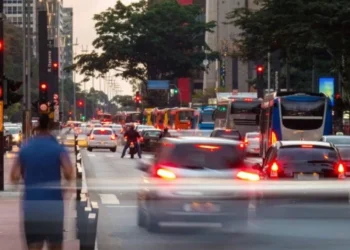 Carros em trânsito com semáforos vermelhos e a presença de veículos de emergência, destacando a importância de dar passagem para ambulância, polícia e bombeiros.
