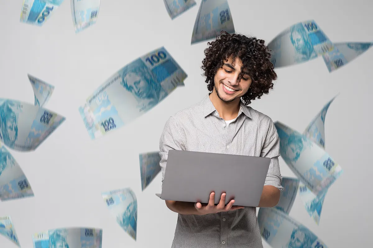 Homem jovem sorridente com cabelo cacheado usando notebook cercado por cédulas de dinheiro voando ao redor