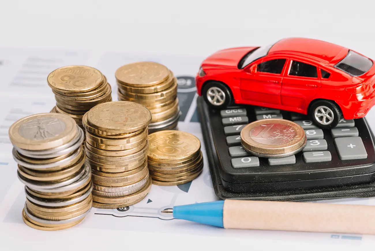 Miniatura de carro vermelho sobre calculadora ao lado de pilhas de moedas e caneta.