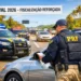 Agente da PRF verifica documentos de motorista em blitz com viaturas e trânsito intenso.