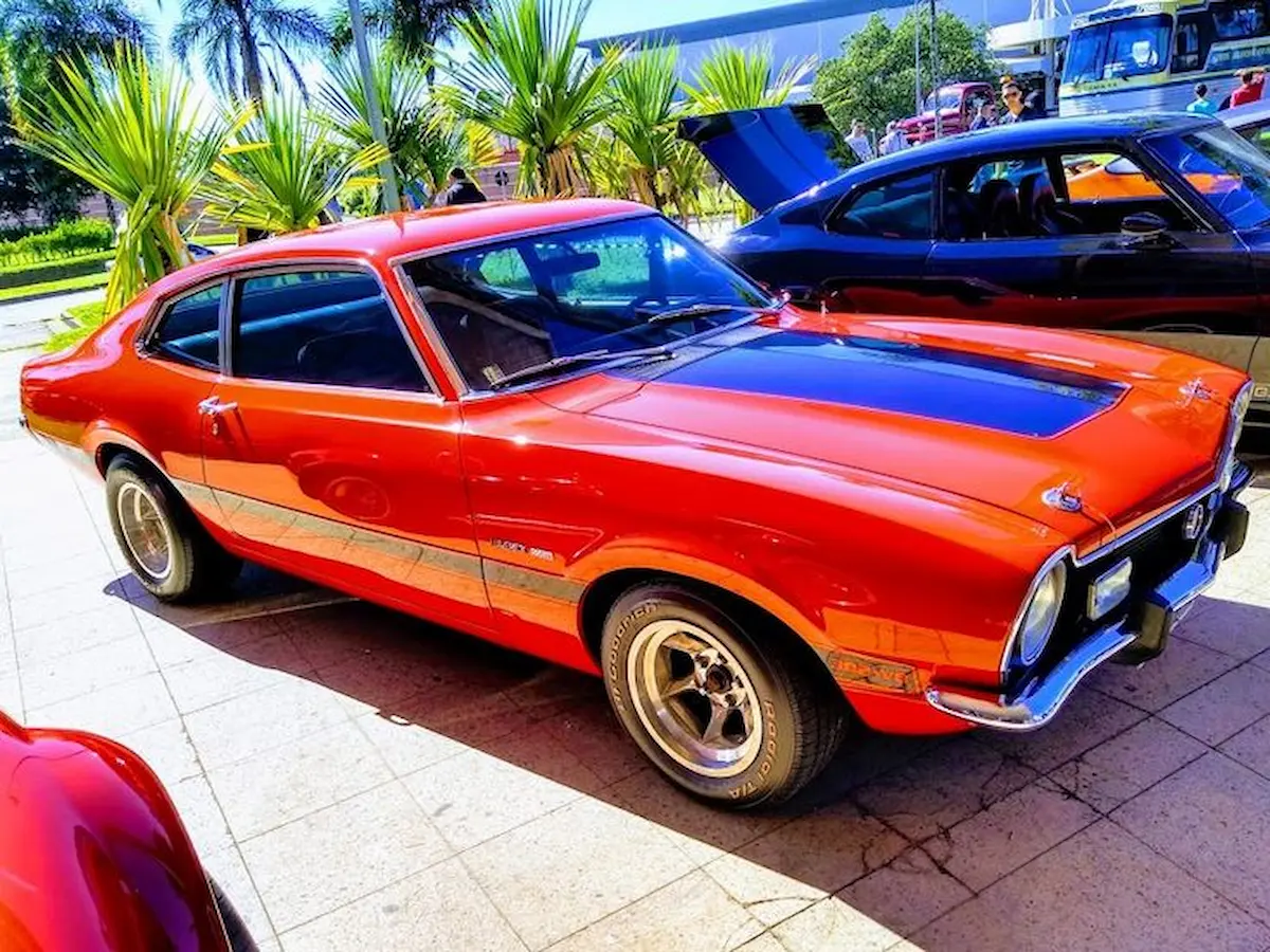 Ford Maverick laranja brilhante estacionado em área externa, cercado por palmeiras e outros carros.