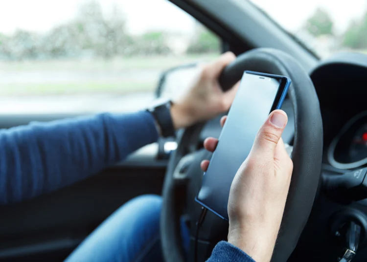 Homem dirige carro enquanto segura celular próximo ao volante na estrada