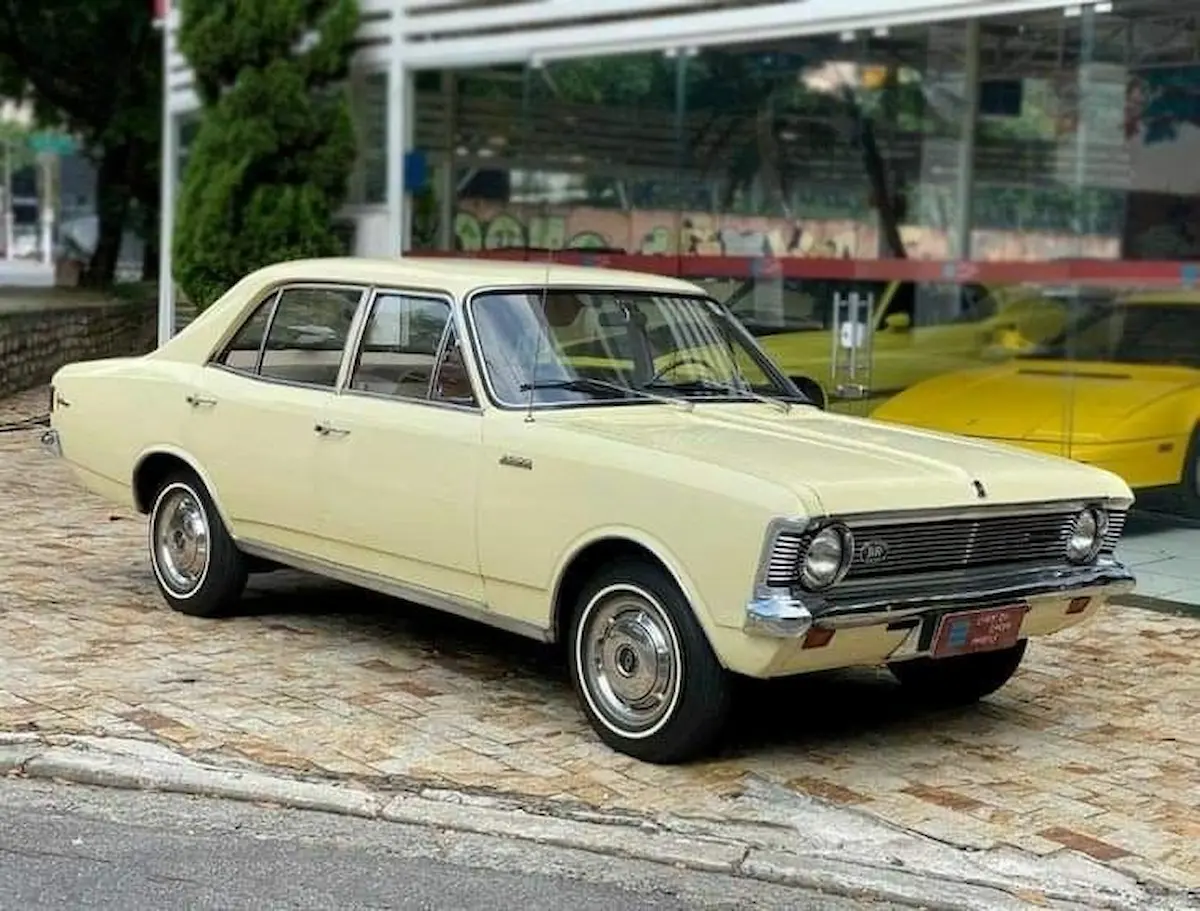 Chevrolet Opala amarelo estacionado em frente a vitrine de concessionária com outro carro antigo ao fundo.