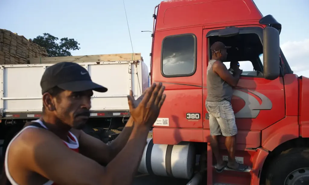 Dois caminhoneiros conversam ao lado e na porta de um caminhão vermelho durante uma parada