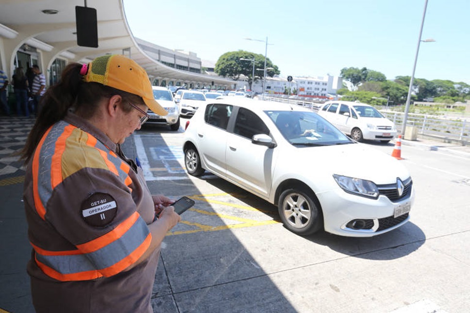 Agente de trânsito registra infração em celular diante de carro branco parado em fila dupla