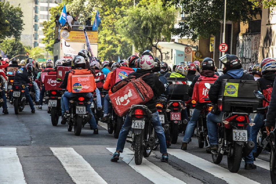 Grupo de motoboys com mochilas de aplicativos parados em faixa de pedestres durante protesto