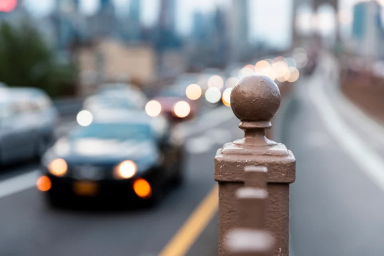 Carros com faróis acesos circulando em via urbana com foco em grade metálica