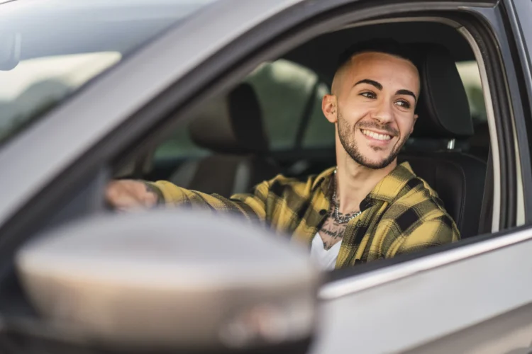 Homem jovem sorridente ao volante de carro visto pelo vidro aberto