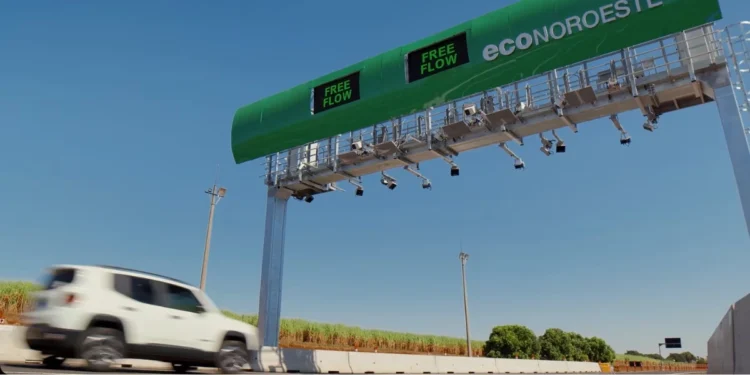 Carro branco passa sob pórtico eletrônico de pedágio em estrada sob céu azul