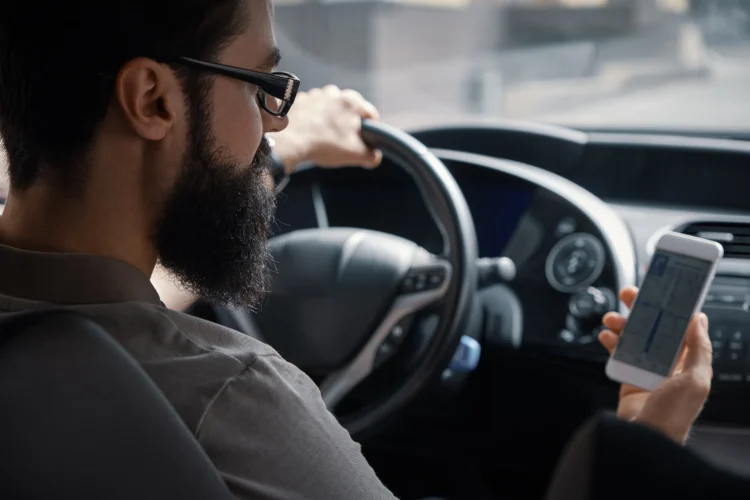 Homem dirigindo carro com uma mão no volante e outra segurando smartphone