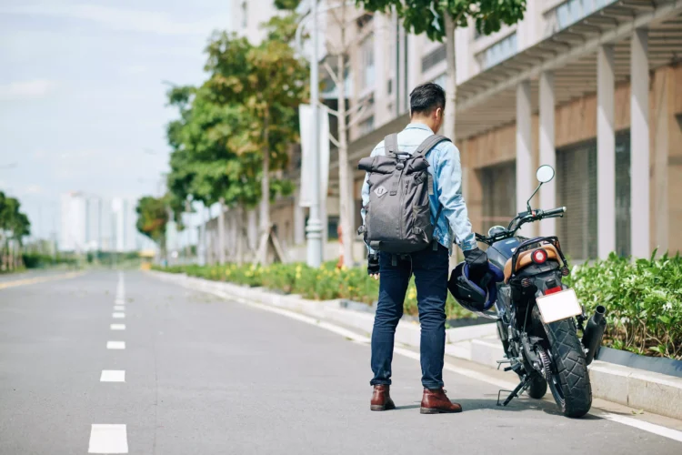 Motociclista com mochila ao lado de moto preta estacionada em rua arborizada