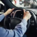Mãos segurando volante de carro, pessoa com camisa azul dirige em ambiente urbano