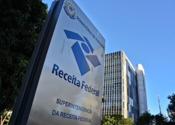 Fachada da Receita Federal com placa azul e logotipo, prédio de vidro ao fundo e céu azul