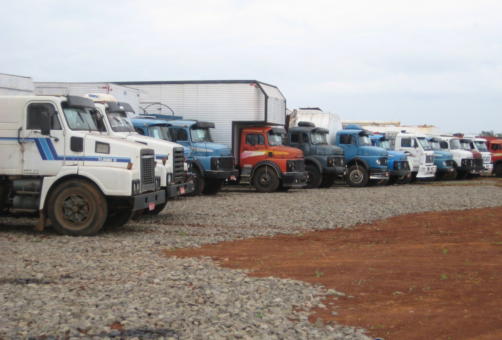 Fileira de caminhões apreendidos em pátio da Receita Federal sobre cascalho, modelos variados em branco, azul e laranja