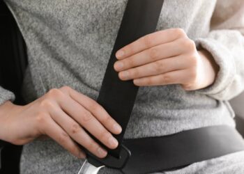 Mãos de pessoa afivelando cinto de segurança preto diagonal cruzando corpo com casaco cinza