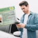 Homem de camisa jeans azul olha celular enquanto CNH brasileira aparece em primeiro plano