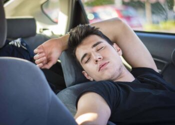 Homem jovem de camiseta preta dorme recostado no banco do passageiro com o braço atrás da cabeça