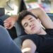 Homem jovem de camiseta preta dorme recostado no banco do passageiro com o braço atrás da cabeça