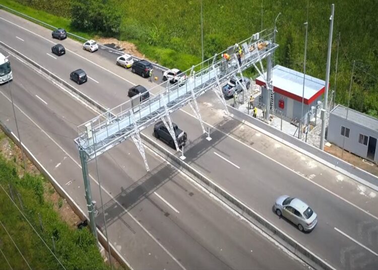 Cobrança eletrônica sobre rodovia, carros passando e técnicos trabalhando ao lado