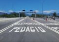 Rodovia com inscrição "PEDÁGIO ELETRÔNICO" no asfalto, pórtico metálico com câmeras e montanhas ao fundo