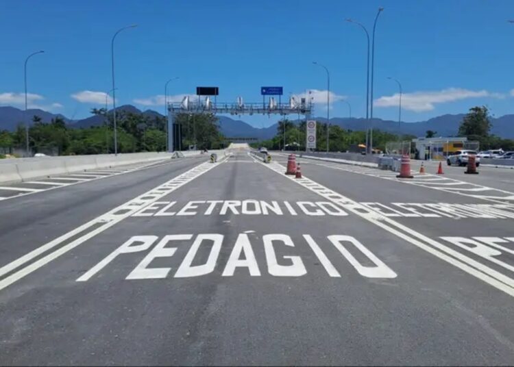Rodovia com inscrição "PEDÁGIO ELETRÔNICO" no asfalto, pórtico metálico com câmeras e montanhas ao fundo