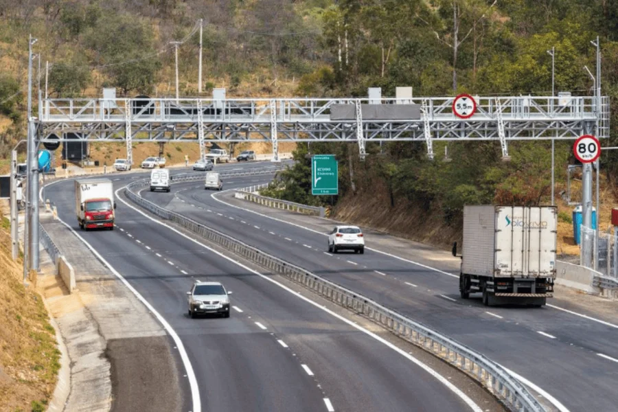 Veículos em rodovia brasileira passando sob pórtico de pedágio eletrônico free flow, com placa de 80 km/h à direita