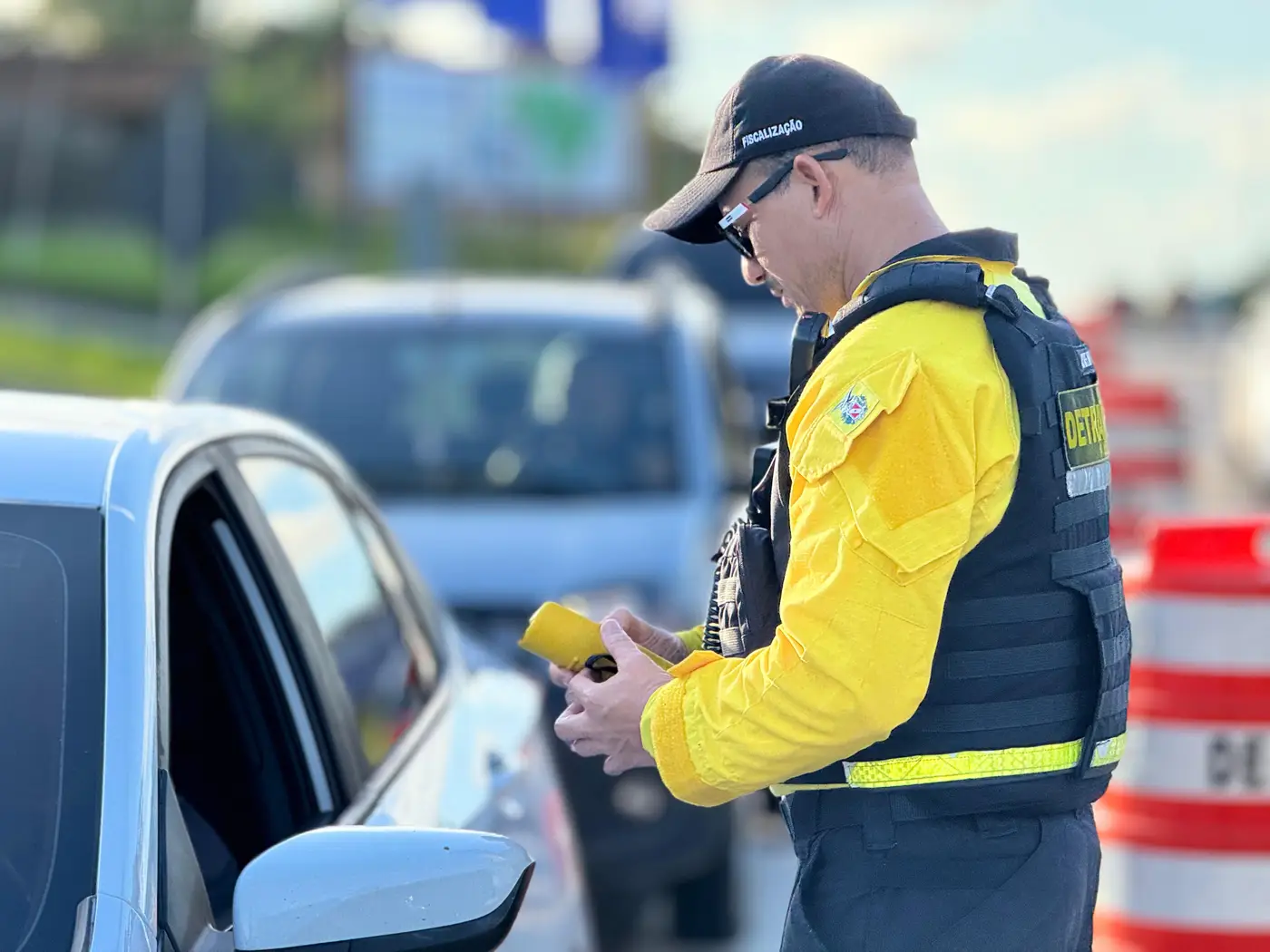 Agente do DETRAN com uniforme amarelo e colete preto verifica documentos de motorista em blitz da Lei Seca