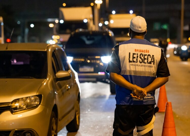 Agente com colete "Operação Lei Seca" observa veículos em blitz noturna com cones laranjas no Rio de Janeiro