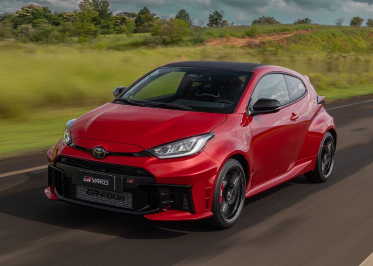 Toyota GR Yaris vermelho com detalhes em preto trafega por estrada asfaltada com paisagem verde ao fundo