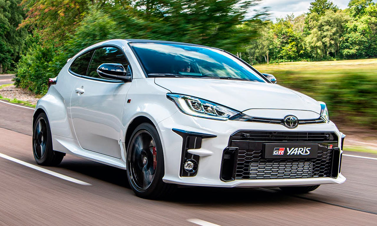 Toyota GR Yaris branco visto de frente em movimento, rodando por estrada arborizada durante o dia