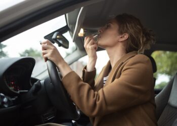 Mulher de blazer marrom passa batom olhando no retrovisor enquanto dirige carro com interior preto