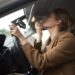 Mulher de blazer marrom passa batom olhando no retrovisor enquanto dirige carro com interior preto