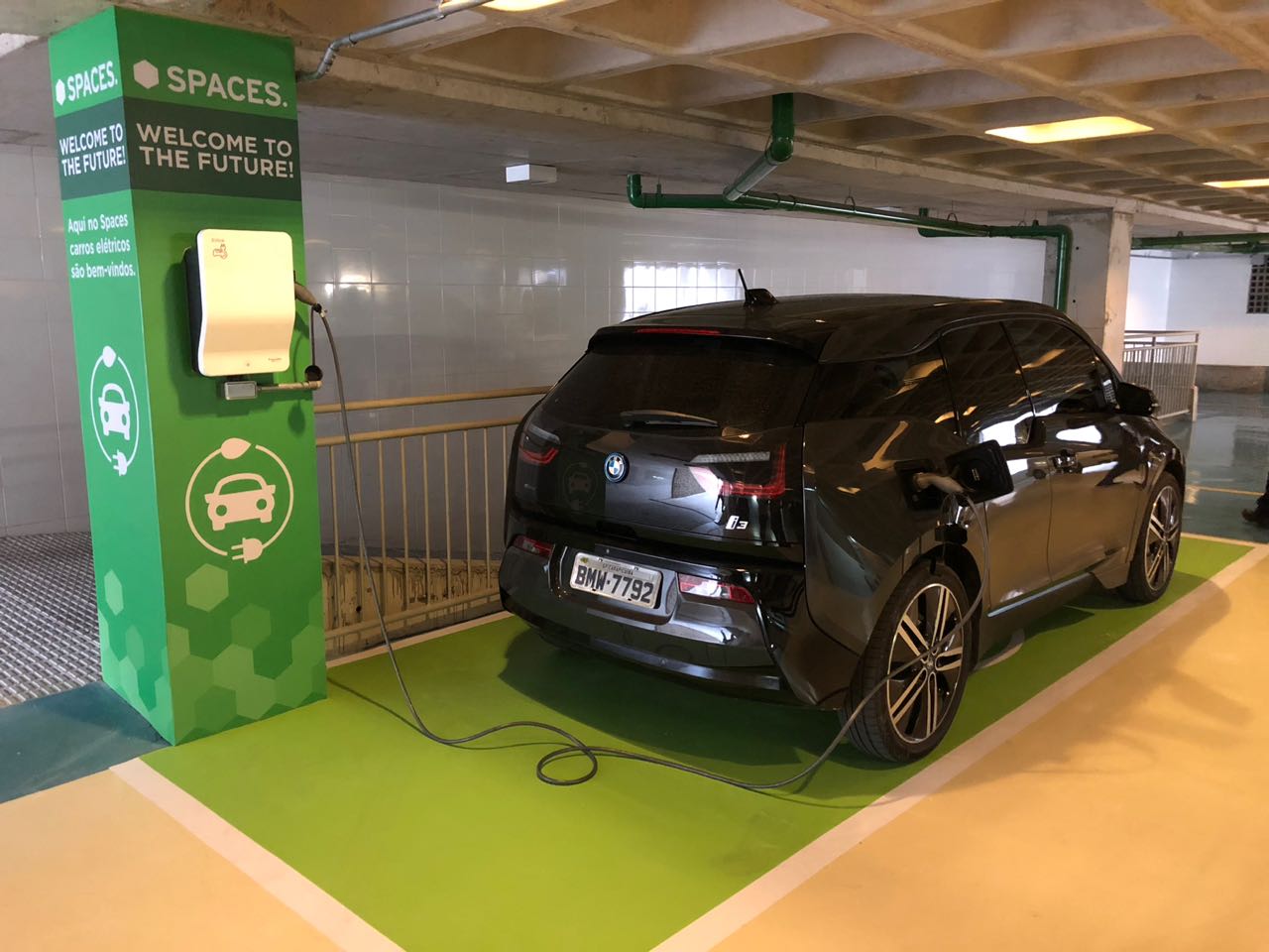 BMW i3 preto conectado a carregador em vaga verde da SPACES, estacionamento com sinalização de veículos elétricos