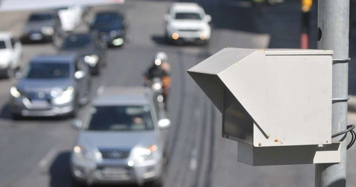 Radar branco fixado em poste monitora fluxo de veículos em via urbana com carros e motocicletas.