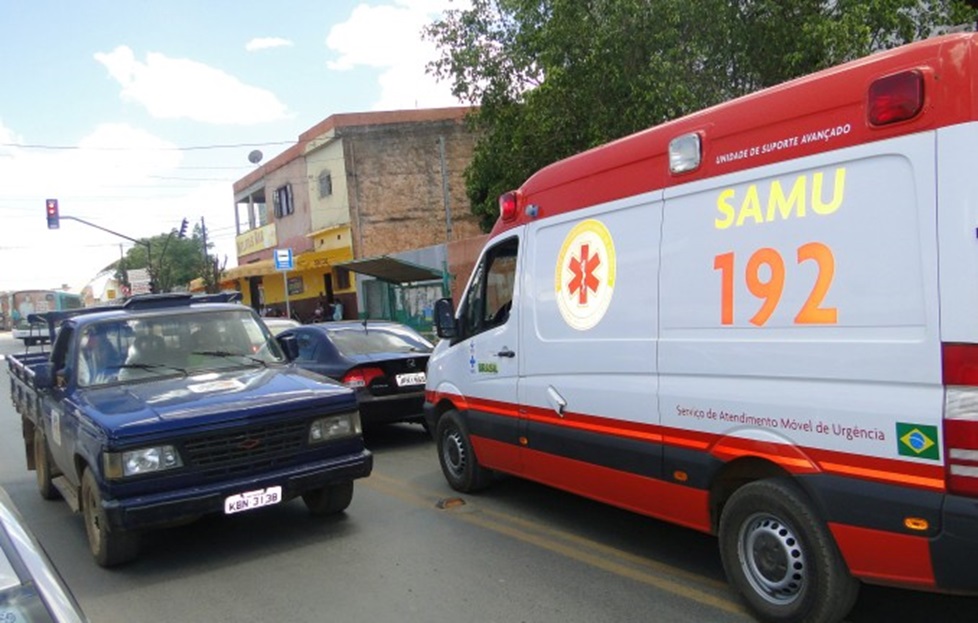 Ambulância branca e vermelha do SAMU 192 parada atrás de caminhonete azul em semáforo de área urbana com casas